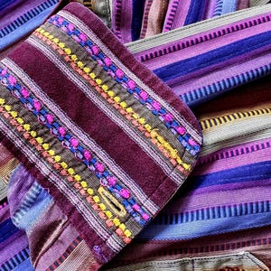 May include: A long-sleeved shirt featuring a vibrant vertical striped pattern in shades of purple, magenta, blue, and tan. The cuff is made of a deep maroon fabric with intricate horizontal embroidery in bright pink, yellow, and blue.