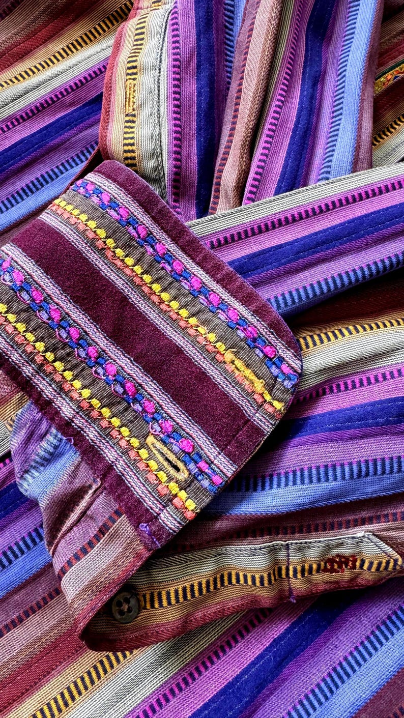 May include: A long-sleeved shirt featuring a vibrant vertical striped pattern in shades of purple, magenta, blue, and tan. The cuff is made of a deep maroon fabric with intricate horizontal embroidery in bright pink, yellow, and blue.