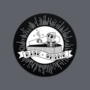 May include: A circular black and white illustration featuring a skeleton in a coffin, holding a coffee cup. The design includes the text "RISE + SHINE" on a banner, surrounded by radiating lines.