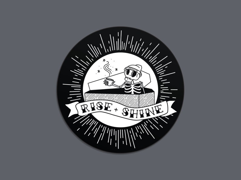 May include: A circular black and white illustration featuring a skeleton in a coffin, holding a coffee cup. The design includes the text "RISE + SHINE" on a banner, surrounded by radiating lines.