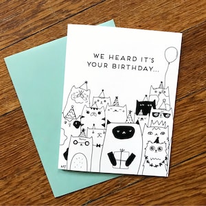May include: A birthday card with a black and white illustration of a group of cats wearing party hats and holding a gift. The text on the card reads "We heard it's your birthday..."