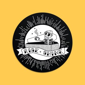 May include: A circular black and white illustration featuring a skeleton in a coffin, drinking coffee. The design includes the text "RISE + SHINE" on a banner, surrounded by radiating lines. The background is yellow.