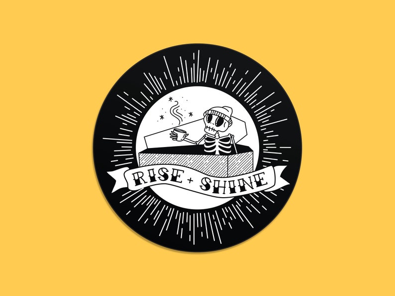 May include: A circular black and white illustration featuring a skeleton in a coffin, drinking coffee. The design includes the text "RISE + SHINE" on a banner, surrounded by radiating lines. The background is yellow.