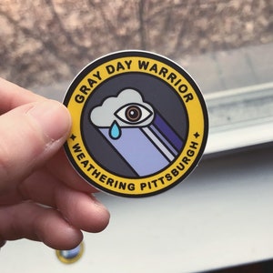 Pu&ograve; includere: Un adesivo circolare giallo e nero con il testo "Gray Day Warrior Weathering Pittsburgh" e un'illustrazione di un occhio che piange dentro una nuvola.