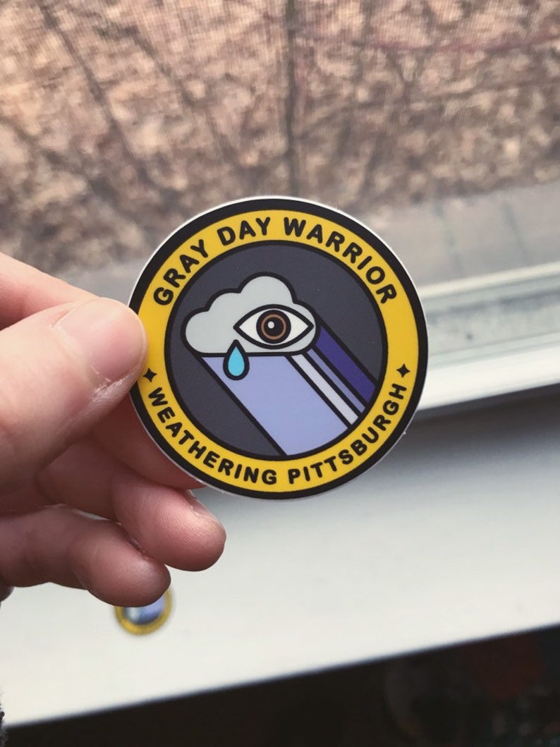 Pu&ograve; includere: Un adesivo circolare giallo e nero con il testo "Gray Day Warrior Weathering Pittsburgh" e un'illustrazione di un occhio che piange dentro una nuvola.