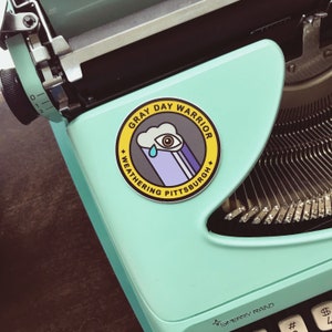 Pu&ograve; includere: Un adesivo giallo e nero con il testo "Gray Day Warrior Weathering Pittsburgh" e un'illustrazione di un cartone animato di una nuvola con un occhio che piange. L'adesivo &egrave; su una macchina da scrivere blu chiaro.