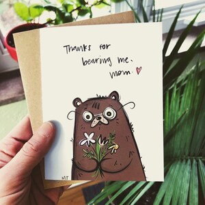 Peut inclure: Carte de v&oelig;ux avec une illustration d'ours brun portant des lunettes et tenant des fleurs. La carte porte l'inscription "Thanks for bearing me, mom." en &eacute;criture noire. La carte est tenue avec une enveloppe marron.