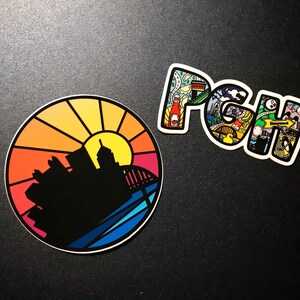 K&ouml;nnte beinhalten: Zwei Aufkleber, einer rund und einer in Form der Buchstaben "PGH". Der runde Aufkleber zeigt einen farbenfrohen Sonnenuntergang &uuml;ber einer Stadtkulisse. Der "PGH"-Aufkleber ist mit bunten Illustrationen von Sehensw&uuml;rdigkeiten in Pittsburgh gef&uuml;llt.