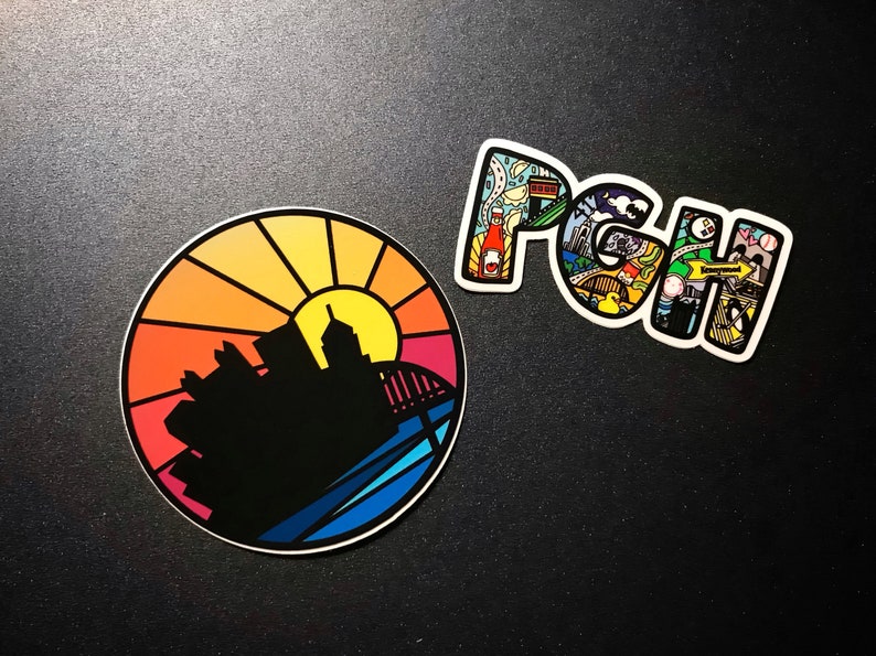 K&ouml;nnte beinhalten: Zwei Aufkleber, einer rund und einer in Form der Buchstaben "PGH". Der runde Aufkleber zeigt einen farbenfrohen Sonnenuntergang &uuml;ber einer Stadtkulisse. Der "PGH"-Aufkleber ist mit bunten Illustrationen von Sehensw&uuml;rdigkeiten in Pittsburgh gef&uuml;llt.