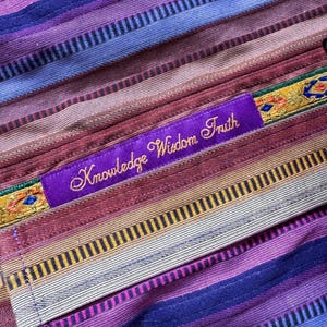 May include: A textile fabric featuring horizontal stripes in shades of purple, blue, red, and tan. A purple rectangular patch is stitched onto the fabric, displaying the embroidered gold text "Knowledge Wisdom Truth" with decorative trim.