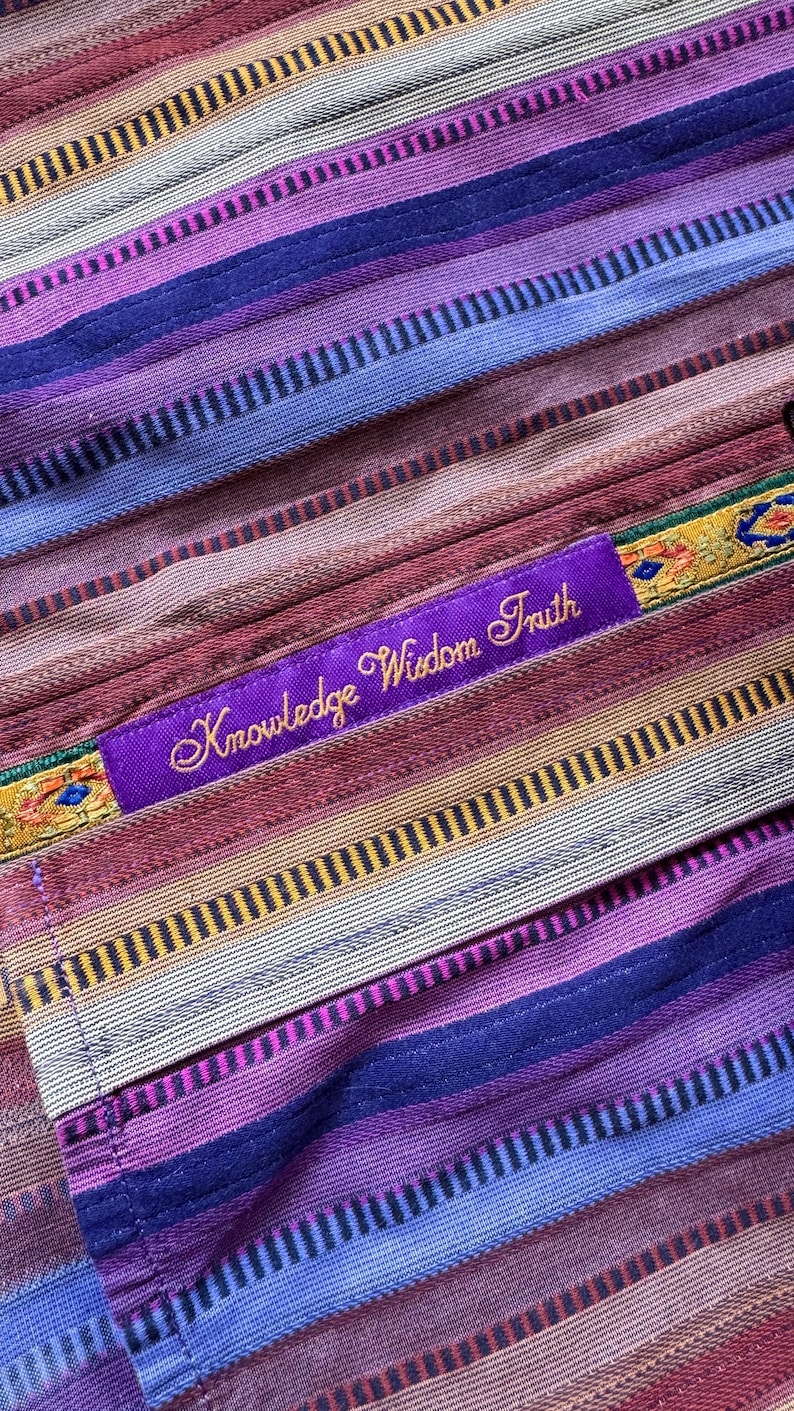 May include: A textile fabric featuring horizontal stripes in shades of purple, blue, red, and tan. A purple rectangular patch is stitched onto the fabric, displaying the embroidered gold text "Knowledge Wisdom Truth" with decorative trim.