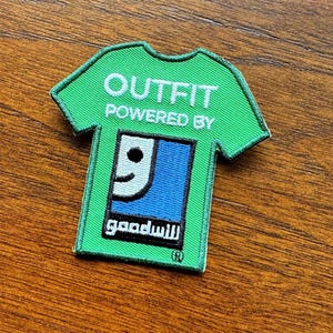Op de afbeelding: Een groene, t-shirtvormige geborduurde patch met witte tekst die OUTFIT POWERED BY leest. Onder de tekst staat een blauw vierkant met een wit cijfer 9 en het woord goodwill in witte kleine letters, met een geregistreerd handelsmerksymbool.