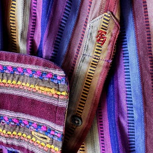 May include: A long-sleeved shirt featuring vertical stripes in shades of purple, blue, red, and tan. The cuff is accented with horizontal bands of pink, blue, and yellow embroidery. A small red embroidered logo is visible on the placket.