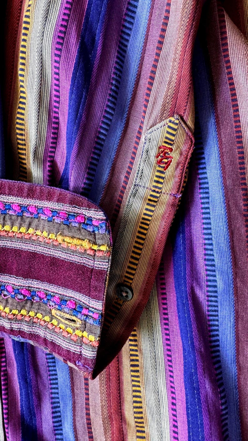May include: A long-sleeved shirt featuring vertical stripes in shades of purple, blue, red, and tan. The cuff is accented with horizontal bands of pink, blue, and yellow embroidery. A small red embroidered logo is visible on the placket.
