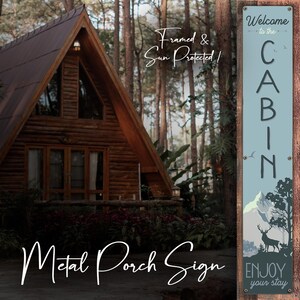 May include: A metal porch sign with the text "Welcome to the CABIN" and a mountain scene with a deer. The sign is framed and sun protected. The sign is in front of a wooden cabin.