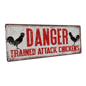 May include: A rustic metal sign with a distressed white background and red text that reads "DANGER TRAINED ATTACK CHICKENS". Two black silhouettes of roosters are on either side of the text.