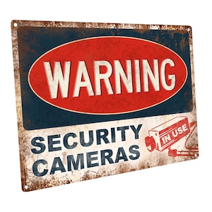 May include: A vintage-style metal sign with a red oval warning sign that says "WARNING" in white letters. Below the warning sign is the text "SECURITY CAMERAS" in black letters. A black and white security camera with the text "IN USE" is in the bottom right corner of the sign.