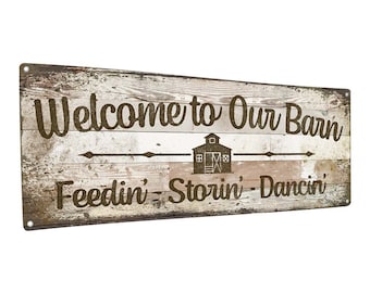 May include: A rustic wooden sign with white paint and brown lettering. The sign reads "Welcome to Our Barn Feedin' Storin' Dancin'". A small barn silhouette is in the center of the sign.