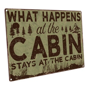 May include: A rustic green metal sign with brown text that reads "What happens at the cabin stays at the cabin". The sign features a silhouette of a mountain, trees, and a sun.