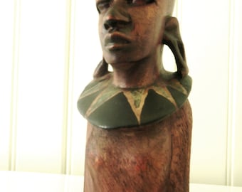 May include: A wooden sculpture of a person's head and shoulders. The sculpture is painted in shades of brown, black, and green. The person is wearing a necklace with a geometric pattern.