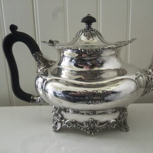 May include: A silver teapot with a black wooden handle and ornate details. The teapot has a round body with a lid and a spout. The base of the teapot has a decorative design.