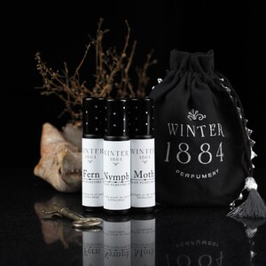 May include: Three small black roll-on perfume bottles with white labels. The labels read "Winter 1884" and the names of the scents: "Fern", "Nymph", and "Moth". The bottles are arranged in a row on a black surface. A black drawstring bag with the text "Winter 1884 Perfumery" is in the background.