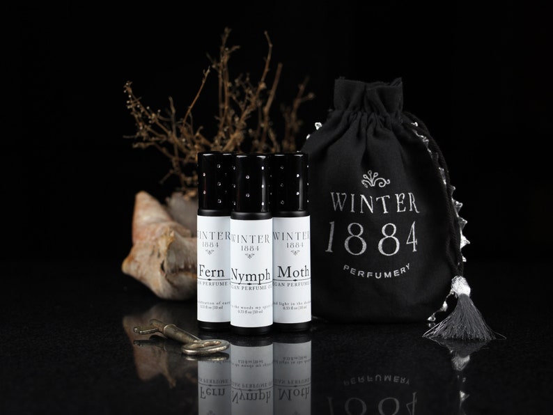 May include: Three small black roll-on perfume bottles with white labels. The labels read "Winter 1884" and the names of the scents: "Fern", "Nymph", and "Moth". The bottles are arranged in a row on a black surface. A black drawstring bag with the text "Winter 1884 Perfumery" is in the background.