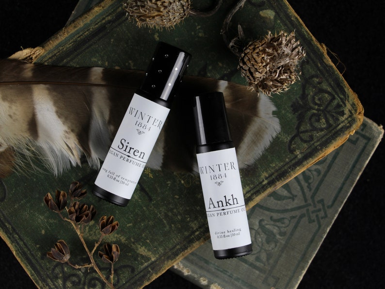 May include: Two small black roll-on bottles with white labels. The labels read "Winter 1884" and "Siren" or "Ankh" with the words "Vegan Perfume Oil" and the size "0.33 fl oz (10 ml)". The bottles are on top of a stack of old, green books with a brown feather and dried flowers.