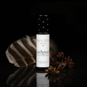May include: A black roll-on bottle of vegan perfume oil labeled "Winter 1881 Kashmer" with the text "The impossible dream 0.33 fl oz / 10 ml" on a black background with a brown and white feather and dried brown flower sprigs.