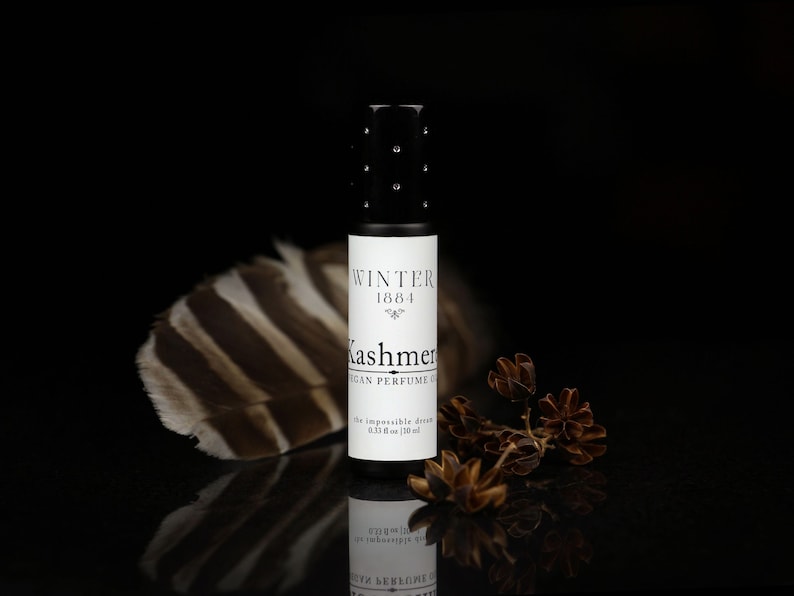 May include: A black roll-on bottle of vegan perfume oil labeled "Winter 1881 Kashmer" with the text "The impossible dream 0.33 fl oz / 10 ml" on a black background with a brown and white feather and dried brown flower sprigs.