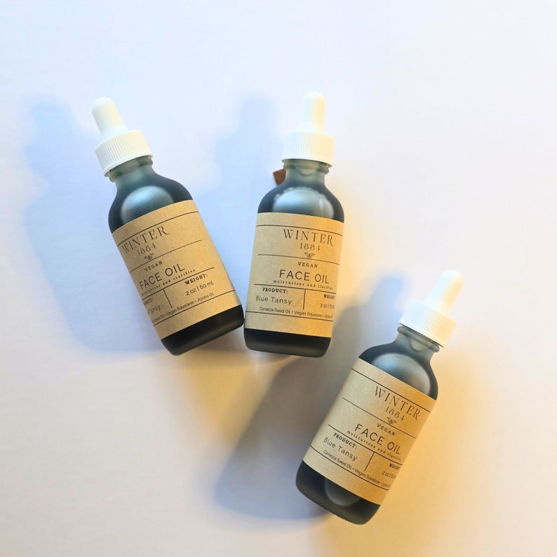 May include: Three glass bottles with dropper tops filled with dark blue liquid. Each bottle has a brown paper label with the text "Winter 1884" and "Vegan Face Oil". The labels also list the product as "Blue Tansy".