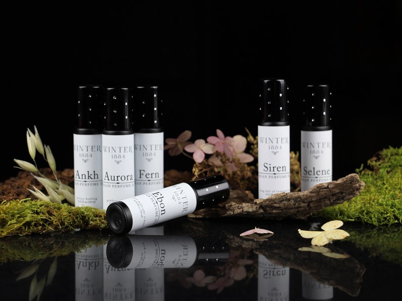 May include: Six small black roll-on bottles with white labels. The labels have the text "Winter 1884" and the names "Ankh", "Aurora", "Fern", "Ebon", "Siren", and "Selene". The bottles are arranged on a wooden surface with moss and flowers.