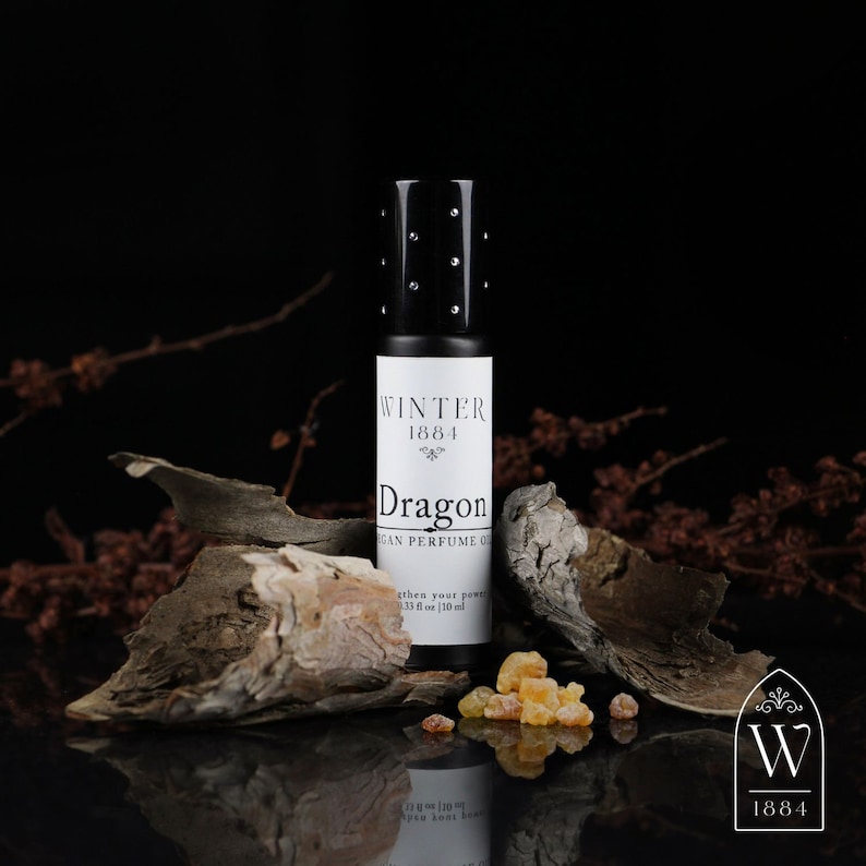 May include: A black roll-on bottle of Dragon vegan perfume oil by Winter 1884. The bottle is sitting on a piece of bark with amber resin crystals scattered around it.