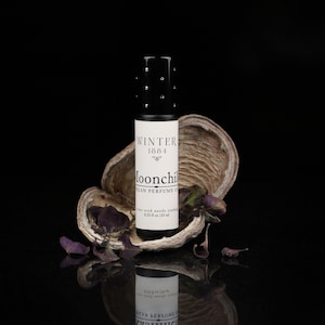 May include: A small black glass bottle with a white label that reads "Winter 1684 Moonchild Vegan Perfume Oil". The bottle is sitting on a piece of dried bark with purple dried leaves.
