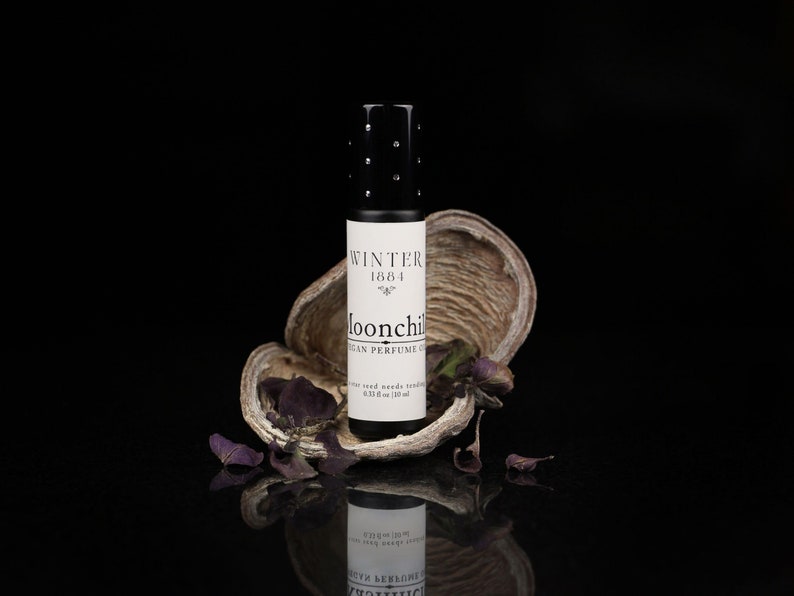May include: A small black glass bottle with a white label that reads "Winter 1684 Moonchild Vegan Perfume Oil". The bottle is sitting on a piece of dried bark with purple dried leaves.