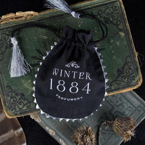 May include: A black velvet pouch with a silver trim and white embroidery. The pouch has the text "Winter 1884 Perfumery" on the front.
