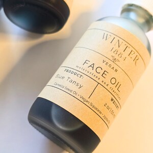 May include: A brown glass bottle with a label that reads "WINTER 1884 VEGAN FACE OIL moisturizes and clarifies PRODUCT: Blue Tansy WEIGHT 2 oz/60ml Camellia Seed Oil + Vegan Squalane + Jojoba Oil"