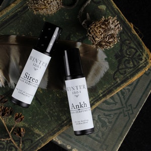 May include: Two small black roll-on bottles with white labels. The labels read "Winter 1881 Siren Vegan Perfume Oil" and "Winter 1884 Ankh Vegan Perfume Oil". The bottles are sitting on top of two old, green books.