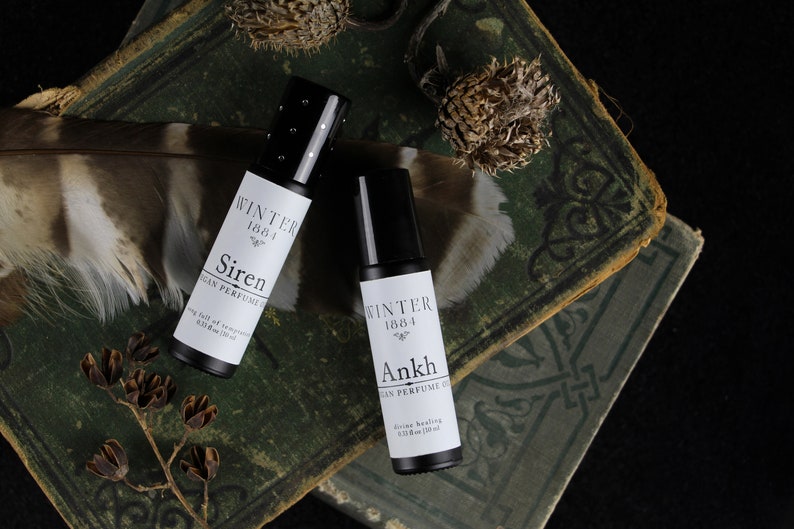 May include: Two small black roll-on bottles with white labels. The labels read "Winter 1881 Siren Vegan Perfume Oil" and "Winter 1884 Ankh Vegan Perfume Oil". The bottles are sitting on top of two old, green books.