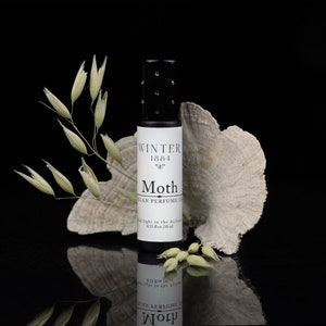 May include: A small glass bottle of vegan perfume called "Moth" by Winter 1864. The bottle is sitting on a white, textured surface with a sprig of dried oats.