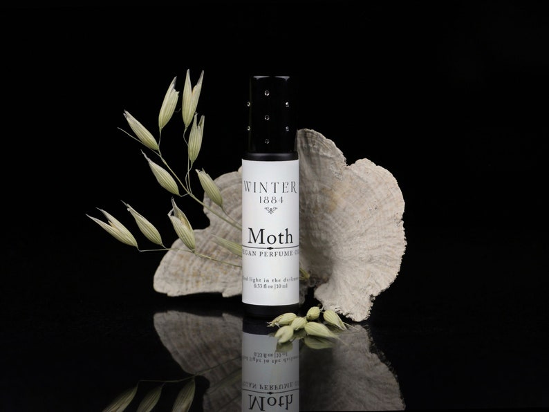 May include: A small glass bottle of vegan perfume called "Moth" by Winter 1864. The bottle is sitting on a white, textured surface with a sprig of dried oats.