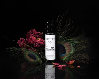 Puede incluir: Un frasco negro con rodillo con una etiqueta blanca que dice "Winter 1881 Sultry Vegan Perfume Oil" y una pluma de pavo real en el fondo. El frasco está rodeado de rosas rojas secas.