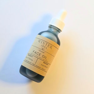 May include: A dark blue glass bottle with a white dropper top and a brown paper label that reads "WINTER 1884 VEGAN FACE OIL moisturizes and clarifies PRODUCT: Blue Tansy WEIGHT: 2 oz / 60 ml Camellia Seed Oil - Vegan Squalane - Jojoba Oil"