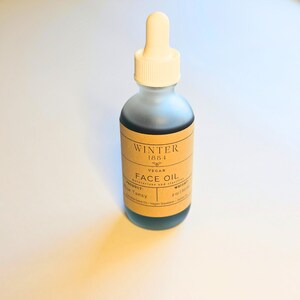 May include: A glass bottle of face oil with a brown label that says "Winter 1884 Vegan Face Oil" and lists the ingredients as "Product: Tansy, Sweet Seed Oil - Vegan Squalane". The bottle is 2 oz and has a dropper.