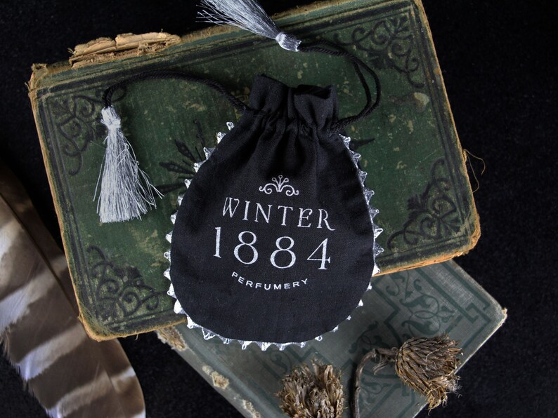 May include: A black velvet pouch with a silver trim and white embroidery. The pouch has the text "Winter 1884 Perfumery" on the front.