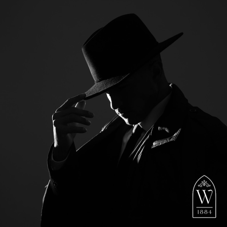 May include: A man in a black fedora and suit jacket, looking down. The image is in black and white and has a vintage feel. The logo "W 1884" is in the bottom right corner.