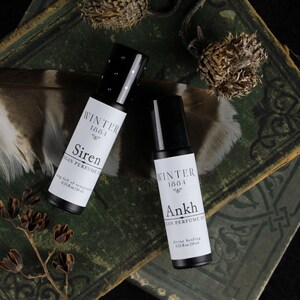 May include: Two small black roll-on bottles with white labels. The labels read "Winter 1884" and "Siren" or "Ankh" with the words "Vegan Perfume Oil" and the size "0.33 fl oz (10 ml)". The bottles are on top of a stack of old, green books with a brown feather and dried flowers.