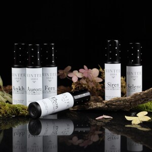 May include: Six small black roll-on bottles with white labels. The labels have the text "Winter 1884" and the names "Ankh", "Aurora", "Fern", "Ebon", "Siren", and "Selene". The bottles are arranged on a wooden surface with moss and flowers.