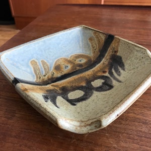 May include: A square ceramic dish with a light blue glaze and brown and black abstract designs. The dish has a slightly raised edge and a rounded lip.