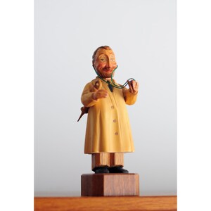 May include: A wooden figurine of a doctor wearing a yellow coat and holding a stethoscope. The figurine is standing on a wooden base.